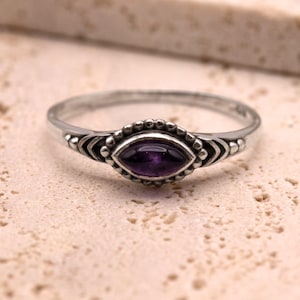 Puede incluir: Un anillo plateado con una piedra preciosa central ovalada de color púrpura oscuro. El anillo presenta un diseño detallado alrededor de la piedra preciosa y a lo largo de la banda, con pequeños detalles de cuentas. El anillo está sobre un fondo neutro.