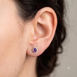 May include: A small, round stud earring featuring a smooth, polished purple gemstone set within a simple silver-toned metal bezel. The earring is worn in the earlobe, showcasing a minimalist design with a vibrant, deep purple centre stone.