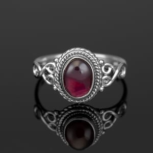 May include: A silver ring featuring a dark red oval gemstone set in a detailed silver bezel. The ring band has a delicate, twisted design.