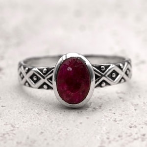 Może przedstawiać: Srebrny pierścionek z owalnym, głębokim, czerwonym kamieniem szlachetnym. Opaska ma powtarzający się geometryczny wzór. Kamień szlachetny jest osadzony w oprawie ramowej, a pierścionek jest prezentowany na teksturowanej powierzchni.