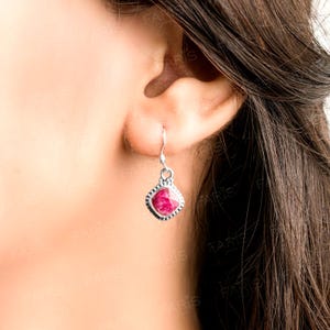 Könnte beinhalten: Ein silberner Ohrring mit einem quadratischen, facettierten, rubinroten Edelstein. Der Ohrring hat eine dekorative Bordüre und einen einfachen Haken. Der Ohrring wird an einem Ohr getragen.