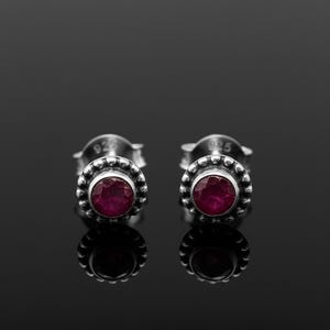 Puede incluir: Un par de pendientes de plata con cierre de tuerca. Cada pendiente presenta una gema redonda de color rojo intenso engastada en un borde con cuentas. Los pendientes se muestran sobre un fondo oscuro, con sus reflejos.