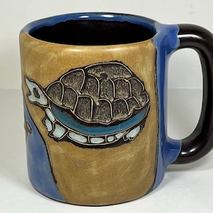 May include: A ceramic coffee mug with a blue and brown glaze. The mug features a hand-painted design of a turtle with a blue shell and white accents.