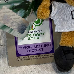 May include: A plush toy mascot from the 2006 FIFA World Cup Germany, wearing a white jersey with the number 06. The toy has yellow fur and black soccer ball-shaped feet. The official licensed product tag is visible.