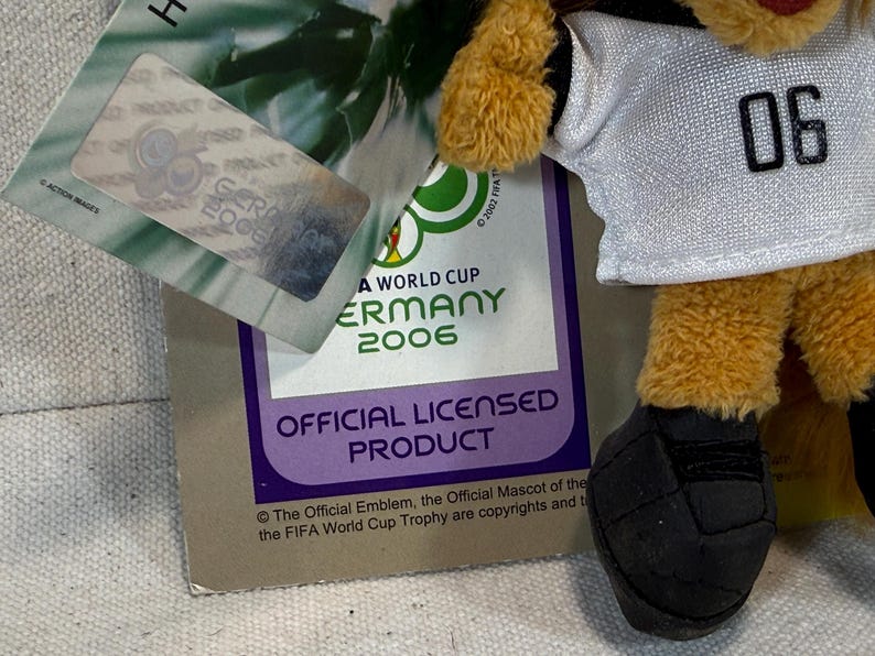 May include: A plush toy mascot from the 2006 FIFA World Cup Germany, wearing a white jersey with the number 06. The toy has yellow fur and black soccer ball-shaped feet. The official licensed product tag is visible.