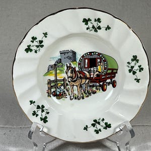 Puede incluir: Un plato de cerámica blanco con borde dorado presenta una ilustración colorida de un carro tirado por un caballo con una lona verde, un castillo en el fondo y diseños de tréboles alrededor del borde.