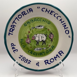 May include: A round ceramic plate with a teal rim, featuring hand-painted text reading TRATTORIA CHECCHINO DAL 1887 ROMA. The center has a green circle with a sheep illustration, two fish, and two pink onions, with the text ABBACCHIO ALLA CACCIATORA.