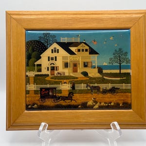 May include: A rectangular ceramic tile framed in light oak wood, featuring a folk art painting of a coastal house with a white picket fence, a horse-drawn carriage labeled 'Breyer', a horse, and a small boat on the water under a blue sky.
