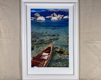 May include: A wooden canoe with a brown and white striped design floats in clear blue water. The canoe is in front of a rocky shoreline with a mountain range in the background.