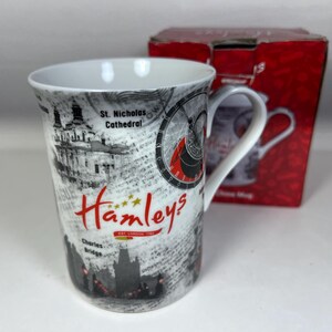 May include: White ceramic mug with a black and white design featuring London landmarks and the text "Hamleys" in red. The mug has a black handle.
