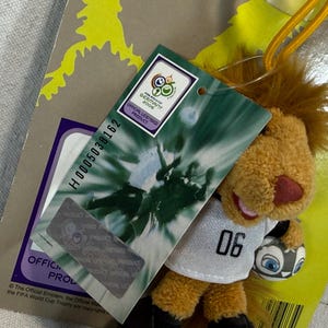 May include: A plush lion toy wearing a white jersey with the number "06" and holding a soccer ball. The toy has a brown mane, a red nose, and is attached to a tag that reads "FIFA World Cup Germany 2006 Official Licensed Product."