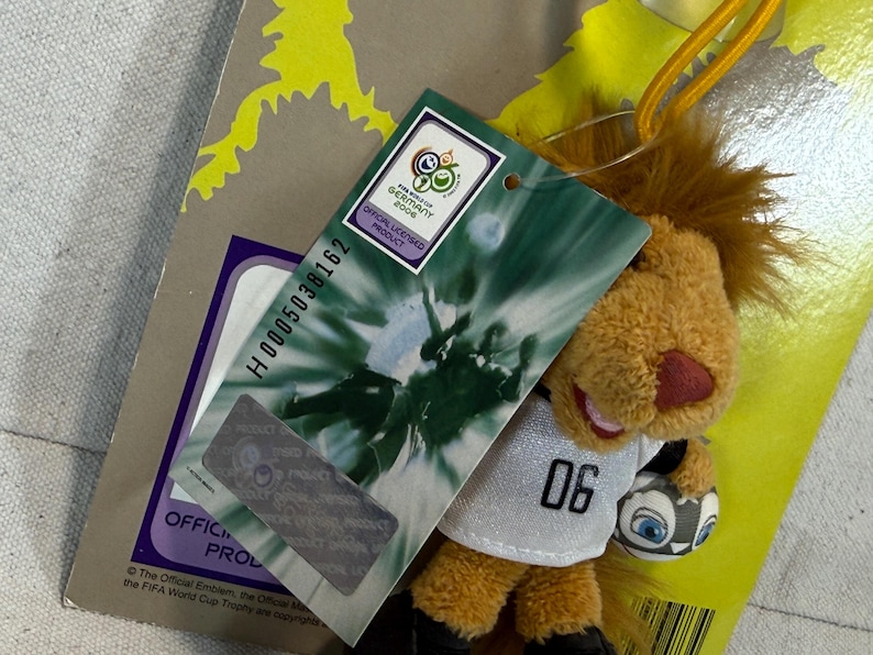 May include: A plush lion toy wearing a white jersey with the number "06" and holding a soccer ball. The toy has a brown mane, a red nose, and is attached to a tag that reads "FIFA World Cup Germany 2006 Official Licensed Product."