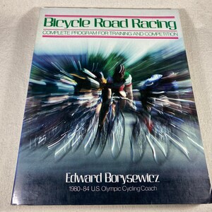 Puede incluir: Una portada de libro con una imagen borrosa de ciclistas corriendo en una carretera. El título es "Bicycle Road Racing: Complete Program for Training and Competition". El autor es Edward Borysewicz, entrenador de ciclismo olímpico de EE. UU. de 1980 a 1984.