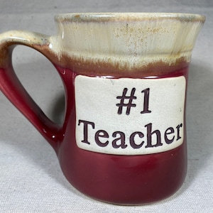 Könnte beinhalten: Eine rote und weiße Keramiktasse mit Henkel. Die Tasse hat ein weißes rechteckiges Etikett mit dem Text "#1 Teacher" in braunen Buchstaben.