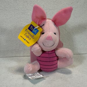 May include: A pink Piglet plush toy with large pink ears, a pink nose, and a striped torso. The toy has a yellow tag that reads "Pooh Friendly Tales" and "Mouse Works". The plush is a children's toy.