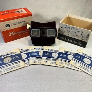 May include: A vintage View-Master 3-D viewer with a brown plastic body and two red viewing lenses. The viewer is surrounded by five View-Master reels, each with the text "One View-Master Reel" and "7 More Wonders of the World".