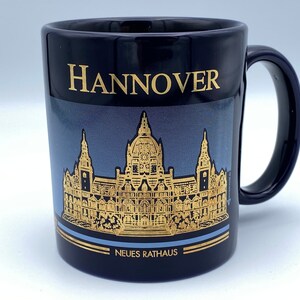 May include: A black ceramic mug with a gold design featuring the Neues Rathaus building in Hannover, Germany. The mug has a gold inscription that reads "Hannover" and "Neues Rathaus".