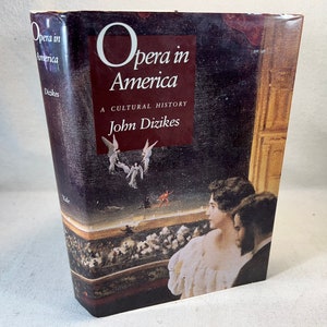Puede incluir: Un libro titulado "Ópera en América: Una historia cultural" de John Dizikes. La portada presenta una pintura de una mujer con un vestido blanco mirando un escenario con ángeles y una figura roja.