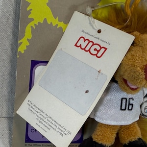 May include: A plush lion toy with a soccer ball, wearing a white jersey with the number "06". The toy has a brown mane, a red nose, and is holding a soccer ball. A tag is attached with the "NICI" logo and text about the FIFA World Cup.