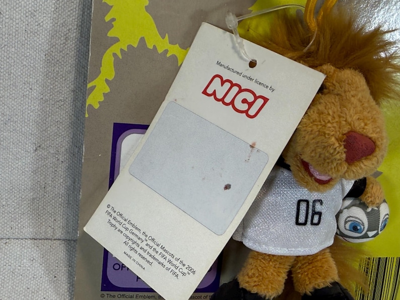 May include: A plush lion toy with a soccer ball, wearing a white jersey with the number "06". The toy has a brown mane, a red nose, and is holding a soccer ball. A tag is attached with the "NICI" logo and text about the FIFA World Cup.