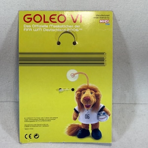 May include: A plush toy of Goleo VI, the official mascot of the 2006 FIFA World Cup Germany, on a yellow cardboard backing. The toy lion wears a white jersey with the number 06 and holds a soccer ball. The packaging includes text in German.
