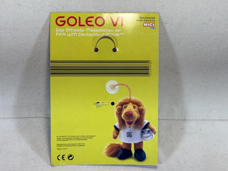 May include: A plush toy of Goleo VI, the official mascot of the 2006 FIFA World Cup Germany, on a yellow cardboard backing. The toy lion wears a white jersey with the number 06 and holds a soccer ball. The packaging includes text in German.