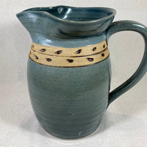 Puede incluir: Una jarra de cerámica azul con una banda marrón y beige alrededor del centro. La jarra tiene una boca ancha y un asa.