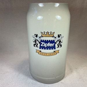 May include: White ceramic beer stein with a blue and gold logo that says "Zipfer" and "SEIT 1858".