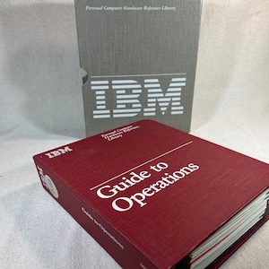 Może przedstawiać: Czerwony segregator z napisem "Guide to Operations" na froncie. Segregator jest oznaczony logo IBM. Segregator leży na szarej książce z logo IBM na froncie.
