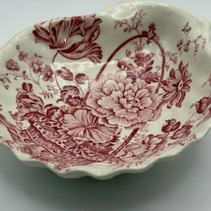 May include: A cream-colored ceramic dish shaped like a shell, featuring a detailed red transferware pattern of a floral basket filled with large blossoms, leaves, and delicate stems. The edges of the dish are scalloped and wavy in design.