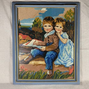 May include: A framed needlepoint picture of two children sitting together. The boy is wearing a brown jacket and blue jeans. The girl is wearing a blue dress. They are both looking at the viewer. The background is a landscape with trees and a house.