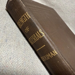 May include: A brown hardcover book with a textured fabric cover. The spine features gold embossed text that reads STRENGTH OF MATERIALS and MERRIMAN. The book is positioned at an angle, showing the front cover and the spine clearly.