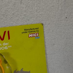 May include: A yellow card with red text that reads "VI" and "ottchen der und 2006". The card also has the text "Manufactured under license by NICI sports". The background is a light gray fabric.