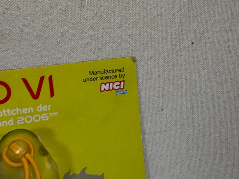 May include: A yellow card with red text that reads "VI" and "ottchen der und 2006". The card also has the text "Manufactured under license by NICI sports". The background is a light gray fabric.