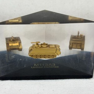May include: A clear acrylic display case containing a gold-coloured M113A3 armoured personnel carrier model. The case has a black base with the text "M113A3 United Defence" printed on it.