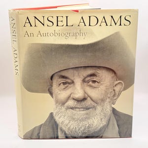 May include: A hardcover book titled Ansel Adams An Autobiography featuring a sepia-toned portrait of a bearded person wearing a wide-brimmed hat on the front cover. The title and author name are printed in black serif font at the top.