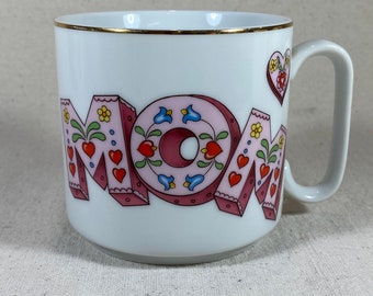 Könnte beinhalten: Weiße Keramiktasse mit goldfarbenem Rand und einem bunten Design des Wortes "MOM" in Rosa, Rot, Grün, Blau und Gelb. Die Buchstaben sind mit Herzen und Blumen verziert.