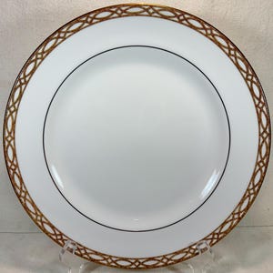 May include: White ceramic plate with a gold and brown geometric border design. The plate has a black inner rim and is displayed on a clear stand. A classic piece for serving food.