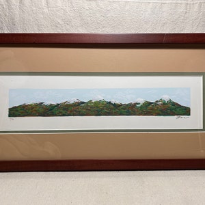 Op de afbeelding: Een ingelijste print van een landschapschilderij dat een bergketen met besneeuwde toppen toont. De bergen zijn groen en bruin met een blauwe lucht en witte wolken op de achtergrond. De print is rechtsonder getekend met "Marino".