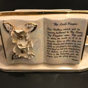 Pode incluir: Um suporte de livros de cerâmica branca em forma de livro aberto com detalhes dourados e florais. O suporte de livros apresenta a Oração do Pai Nosso impressa na frente.