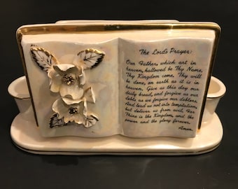 May include: A white ceramic bookend shaped like an open book with gold trim and floral accents. The bookend features the Lord's Prayer printed on the front.