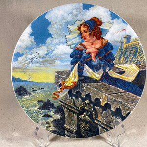 Puede incluir: Un plato de cerámica con una imagen pintada de una mujer con un vestido azul y velo blanco de pie en un borde de piedra con vistas a una masa de agua. La mujer mira hacia el mar y hay pájaros volando en el cielo. El plato está firmado "Haworth" en la esquina inferior derecha.