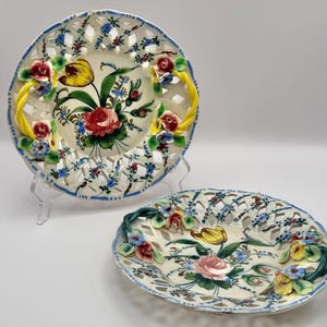 May include: Two round ceramic plates with openwork lattice borders and scalloped edges. The centers feature hand-painted floral motifs including red roses and yellow tulips. Each plate has three-dimensional ceramic flowers and twisted handles.