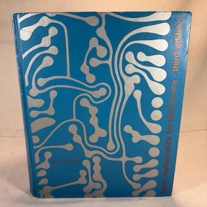 Puede incluir: Un libro de tapa dura azul con un diseño abstracto plateado en la portada. El título es "basic mathematics for electronics - third edition". Se trata de un libro de texto, probablemente para fines educativos.