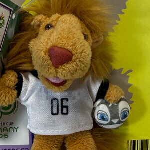 May include: A plush lion toy wearing a white jersey with the number "06" and holding a soccer ball. The lion has a red nose and a smiling expression. The toy is an official licensed product from the 2006 World Cup in Germany.