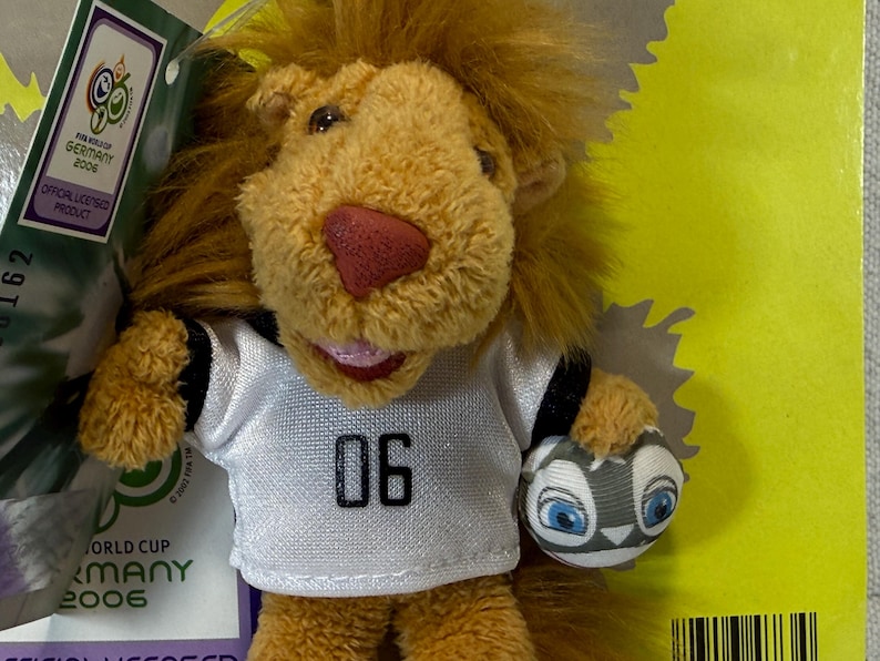 May include: A plush lion toy wearing a white jersey with the number "06" and holding a soccer ball. The lion has a red nose and a smiling expression. The toy is an official licensed product from the 2006 World Cup in Germany.