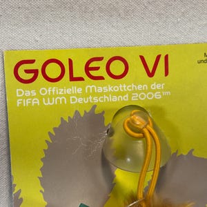 May include: A yellow tag with the text "GOLEO VI" in red. Below, it reads "Das Offizielle Maskottchen der FIFA WM Deutschland 2006". A small, clear plastic charm with a yellow string is attached. The tag is manufactured under license by NICI.