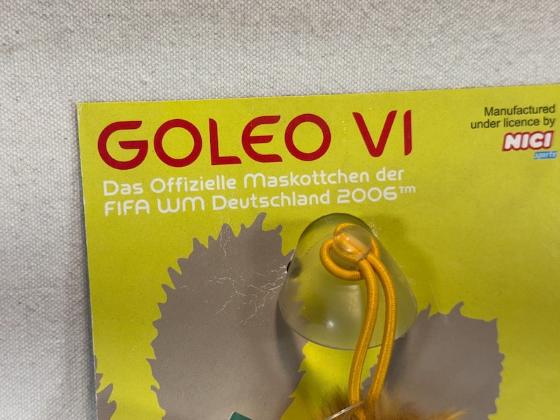 May include: A yellow tag with the text "GOLEO VI" in red. Below, it reads "Das Offizielle Maskottchen der FIFA WM Deutschland 2006". A small, clear plastic charm with a yellow string is attached. The tag is manufactured under license by NICI.