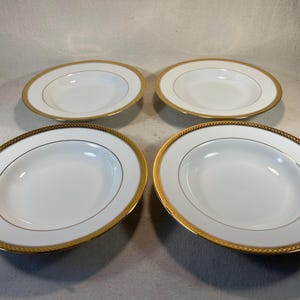 May include: Four white soup bowls with gold trim. The bowls are arranged in a square pattern on a white surface.