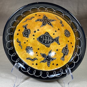 May include: A decorative ceramic plate with a black rim and a yellow center. The plate features black illustrations of fish, starfish, and other sea creatures. A white swirl design decorates the rim.
